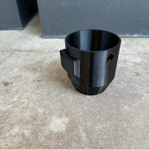 May include: A black plastic cup with a small black handle on the side. The cup is sitting on a concrete surface.