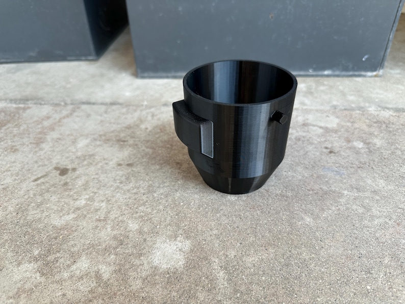 May include: A black plastic cup with a small black handle on the side. The cup is sitting on a concrete surface.