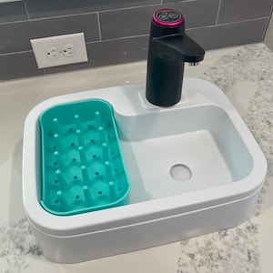 Può includere: Un bacino rettangolare bianco con bordi arrotondati, dotato di un inserto in silicone turchese con pioli in rilievo sul lato sinistro e un distributore d'acqua automatico nero con un anello magenta sul lato destro, su un piano bianco.