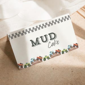 May include: A rectangular card with the words "MUD Cake" in a playful font. The card is decorated with a checkered border and a row of colorful monster trucks. The card is on a light brown surface.