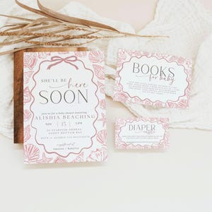May include: A baby shower invitation suite with a seashell theme. The main invitation reads "She'll be here soon" and includes details for the event. Additional cards include "Books for baby" and "Diaper raffle". The design is in shades of pink.