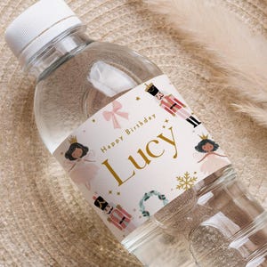 May include: A clear plastic water bottle with a white cap. The label reads "Happy Birthday Lucy" with illustrations of a ballerina, a nutcracker, and a snowflake. The label has a pink background with gold accents. The bottle is partially filled with water.