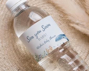 Op de afbeelding: Een doorzichtige plastic waterfles met een witte dop en een etiket met de tekst "Sea you Soon BABY! Aleshia's Baby Shower". Het etiket toont een blauwe walvisillustratie en aquatische plantenontwerpen. De fles is gedeeltelijk gevuld met water.