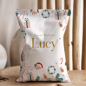 May include: A white gift bag with a festive Christmas design, featuring Santa Claus, presents, and candy canes. The words "Happy Birthday Lucy" are printed in gold. The bag is sitting on a wooden surface.