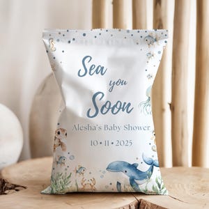 May include: A white baby shower favor bag with a sea-themed design. The bag features illustrations of sea creatures, including a whale, turtle, and fish, with the text "Sea you Soon" and the baby shower details.