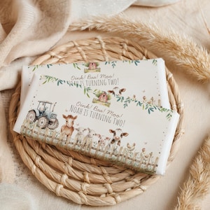 May include: Two white candy bar wrappers with a farm animal theme. The wrappers feature a tractor, cows, sheep, chickens, and the text "Oink! Baa! Moo! Noah is turning two!" on a wooden fence background.