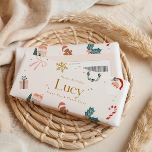Może przedstawiać: Prezent zapakowany w kremowy papier z świątecznymi ilustracjami domów, piernikowych ludzików i Świętego Mikołaja. Tekst "Happy Birthday Lucy" jest w kolorze złotym. Prezent umieszczono na plecionej macie z wikliny.