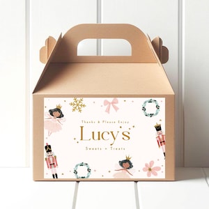 Könnte beinhalten: Eine braune Geschenkbox aus Pappe mit Griff und rechteckigem Etikett. Das Etikett zeigt den Text "Lucy's Sweets + Treats" mit Illustrationen einer Fee, eines Nussknackers, einer Schneeflocke, einer Schleife, eines Kranzes und einer Blume. Der Text "Thanks & Please Enjoy" ist ebenfalls vorhanden.