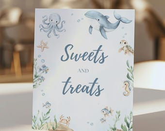 May include: A white sign with the words "Sweets and Treats" in blue script, decorated with watercolor illustrations of sea creatures. The design includes an octopus, whale, starfish, fish, crab, seahorse, and turtle. The sign is held by a gold metal stand.