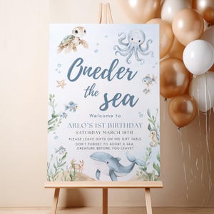 May include: A welcome sign for a first birthday party, featuring an "Under the Sea" theme. The sign includes illustrations of sea creatures like a turtle, octopus, and whale, with the text "Oneder the Sea". The sign is displayed on an easel.