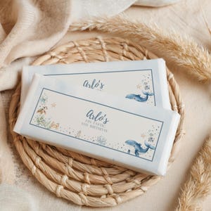 May include: Two wrapped candy bars with a light blue border and aquatic illustrations. The text "Arlo's" and "Fin-tastic 5th Birthday" are printed on the front. The bars are resting on a woven mat with dried grass.