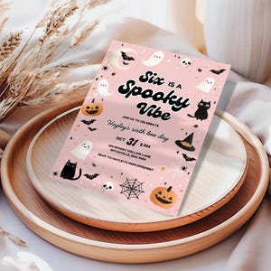 Peut inclure: Une invitation rose sur le th&egrave;me d'Halloween avec le texte "Six is a Spooky Vibe". L'invitation comprend des illustrations de fant&ocirc;mes, de chauves-souris, de citrouilles et un chapeau de sorci&egrave;re. L'invitation est sur un plateau en bois.