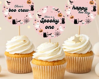 Op de afbeelding: Drie cupcakes met witte frosting en decoratieve toppers. De toppers zijn roze cirkels met Halloween-thema illustraties en tekst die luidt "Olivia's boo crew", "Spooky one" en "happy boo day".
