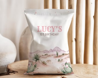 Puede incluir: Una bolsa de recuerdo de fiesta blanca con el texto "LUCY'S 1ST BIRTHDAY" en rosa. La bolsa presenta un diseño de paisaje desértico con cactus, montañas y una paleta de colores apagados. La bolsa está sobre una superficie de madera.