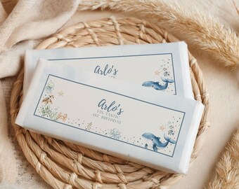 May include: Two wrapped candy bars with light blue borders and the text "Arlo's FIN-tastic 1st Birthday." The bars feature illustrations of whales, starfish, and other sea creatures. The bars are on a woven mat.