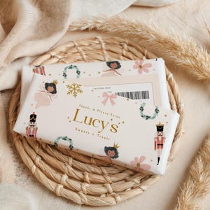 May include: Two wrapped chocolate bars with a festive design featuring fairies, nutcrackers, and snowflakes. The bars are labeled "Lucy's Sweets + Treats" in gold lettering. The bars rest on a woven wicker placemat.