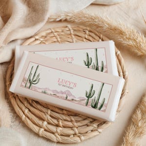 May include: Two chocolate bars with a desert theme. The bars feature a light pink background with a watercolour illustration of cacti and a desert landscape. The text "LUCY'S 1ST BIRTHDAY" is printed in pink. The bars are on a woven basket.