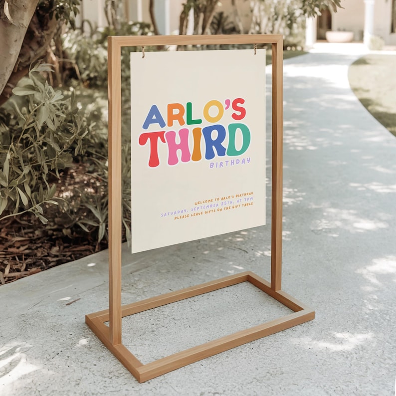 May include: A rectangular sign suspended from a light wood frame. The sign features the colourful text ARLO'S THIRD BIRTHDAY in a retro font, with additional smaller blue text providing event details including the date, time, and gift instructions.