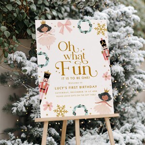 Pode incluir: Uma placa branca em um cavalete de madeira com o texto "Oh what Fun, It is to be One!" e "Lucy's First Birthday". A placa é decorada com ilustrações de fadas, quebra-nozes, flocos de neve e grinaldas. O fundo é uma cena de inverno nevada.