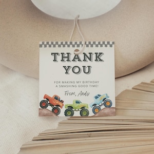 以下が含まれることがあります： チェック柄のボーダーが付いた正方形のギフトタグ。タグには太字のグレーの文字で「THANK YOU」と書かれています。その下に「FOR MAKING MY BIRTHDAY A SMASHING GOOD TIME! From, Andy.」と書かれています。3台のカラフルなモンスタートラックが下にあります。