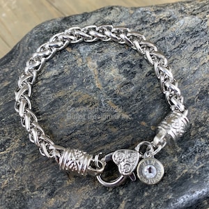 May include: A silver chain bracelet with a heart-shaped clasp and a small charm with a clear crystal. The bracelet is made of braided metal and has a decorative end cap.