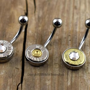 May include: Three silver belly button rings with different bullet casings. The first ring has a clear crystal in the center of the casing. The second ring has a gold-colored casing. The third ring has a clear crystal in the center of the casing.