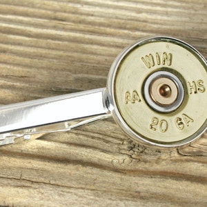 May include: A silver tie clip with a gold-colored shotgun shell as the decorative element. The shell is marked with "WIN AA 20 GA HS".