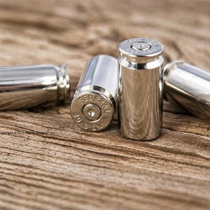 May include: Four polished silver bullet casings are arranged on a weathered wooden surface. The casings have a reflective sheen, with one displaying the text "FEDERAL" and other markings. The image is a close-up, highlighting the metallic texture.