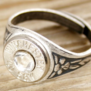 May include: A silver ring with a bullet casing design. The ring features a clear crystal stone set in the center of the bullet casing. The band of the ring is engraved with a floral design.