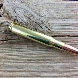 May include: A gold-colored bullet casing keychain with a silver key ring. The casing is open at the top and has a copper-colored tip. The keychain is lying on a wooden surface.
