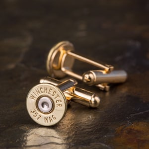 May include: Gold cufflinks featuring a .357 Winchester bullet casing. The cufflinks are made of metal and have a polished finish.