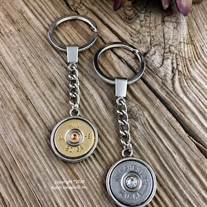May include: Two keychains with silver-tone chains and key rings. One keychain has a gold-tone bullet casing with "AA HS 20 GA" text. The other has a silver-tone bullet casing with "WINCHESTER 20 GA" text and a crystal. Copyright 2026 Bullet Designs Inc.