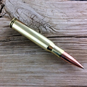 May include: A gold-colored bullet casing pen with a copper tip. The pen is lying on a wooden surface.