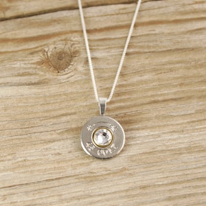 May include: A silver necklace with a round pendant featuring a clear rhinestone set in a silver casing. The pendant is a repurposed bullet casing with the inscription "45 AUTO" visible.