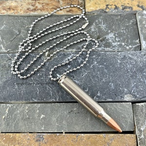 May include: A silver chain necklace with a bullet casing pendant. The bullet casing is silver with a copper tip.