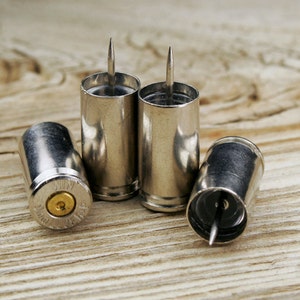 May include: Four silver bullet casings with a small pin protruding from the top of each. The casings are arranged in a row on a wooden surface.