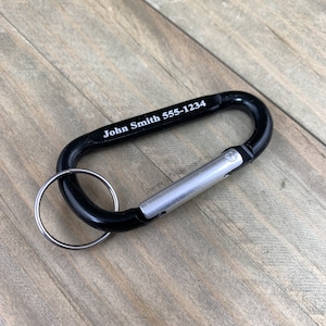 May include: Black carabiner keychain with a silver clip and a silver ring. The keychain is engraved with the text "John Smith 555-1234".