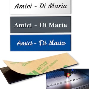 Può includere: Tre targhette rettangolari bianche, grigie e blu, ciascuna con il testo "Amici - Di Maria" in corsivo. Un cuscinetto adesivo nero con uno strato protettivo è visibile sotto. L'immagine mostra anche l'incisione laser.
