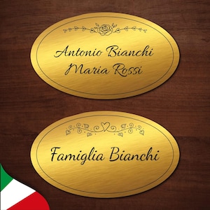Può includere: Due targhe ovali dorate con scritte nere e disegni floreali decorativi. La targa superiore recita "Antonio Bianchi Maria Rossi" e la targa inferiore recita "Famiglia Bianchi".