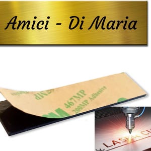 Può includere: Un'insegna rettangolare color oro con le parole "Amici - Di Maria" in corsivo nero. L'insegna è mostrata con un cuscinetto adesivo nero e un primo piano di una macchina da taglio laser.