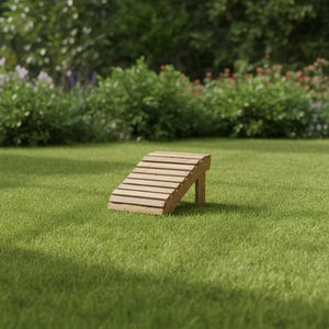 May include: A wooden footrest with a slatted top and a single supporting leg. The footrest is made of light-colored wood and is angled for comfort. It is set on a green lawn with a blurred background of trees and foliage.