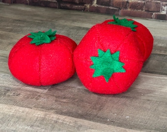Può includere: Quattro ornamenti di pomodoro in feltro rosso con foglie in feltro verde. I pomodori sono disposti in fila su una superficie di legno.