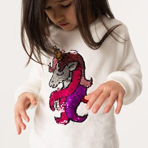 Pode incluir: Camisola branca de manga comprida com um design de unicórnio com lantejoulas em tons de rosa, roxo e prata. O unicórnio tem um chifre dourado. A camisola tem punhos canelados e gola redonda.