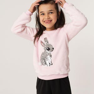 Puede incluir: Sudadera rosa claro y difusa con un diseño de conejo con lentejuelas. El modelo lleva una diadema de orejas de conejo blancas. El conjunto se completa con pantalones negros. Ideal para Pascua o eventos temáticos.