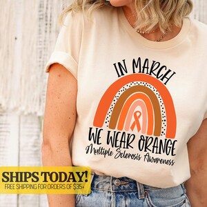 May include: Cream-colored t-shirt with the text "IN MARCH WE WEAR ORANGE" above a rainbow graphic. The graphic includes an orange ribbon, symbolizing Multiple Sclerosis Awareness.