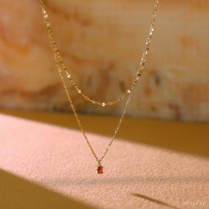 Op de afbeelding: Een goudkleurige ketting met twee lagen en een kleine, ronde, oranje edelsteenhanger. De ketting heeft twee strengen, een met kleine, platte, cirkelvormige schakels en de andere met een fijne ketting. De achtergrond is een zachte, neutrale kleur.