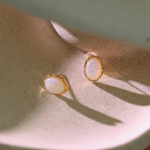 Op de afbeelding: Een paar gouden oorstekers met een ovale, iriserende opaal in het midden. De oorbellen hebben een getextureerde gouden rand en worden gepresenteerd op een neutrale ondergrond. Een sieraad.