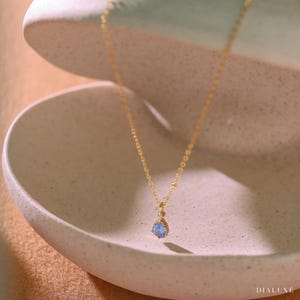 Op de afbeelding: Een delicate gouden ketting met een druppelvormige, lichtblauwe edelsteen hanger. De hanger is gezet in een gouden frame en hangt aan een fijne gouden ketting. De ketting wordt gepresenteerd in een lichtgekleurde, getextureerde kom.