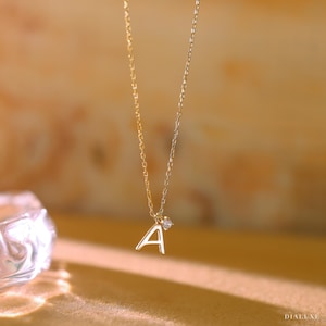 Peut inclure: Un délicat collier en or avec un pendentif en forme de lettre "A" et un petit diamant. La chaîne est fine et élégante, avec une teinte dorée chaleureuse. Le collier est présenté sur un fond flou.