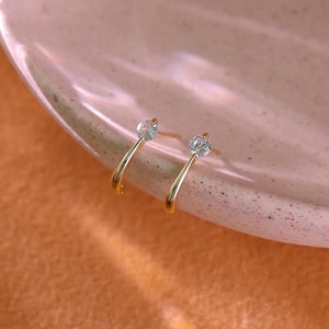 May include: A pair of gold-coloured hoop earrings, each with a clear, round gemstone at the top of a curved post. The earrings are presented on a light-coloured, speckled surface.